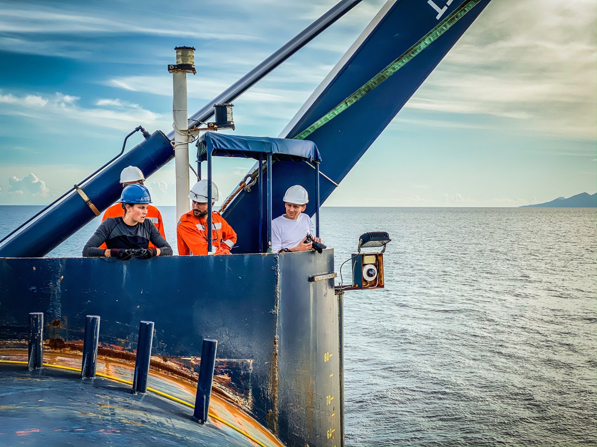 Four members of the expedition team wearing hard hats, lean against the wall of the ship's deck watching the deployment of the ROV Nereid Under Ice.