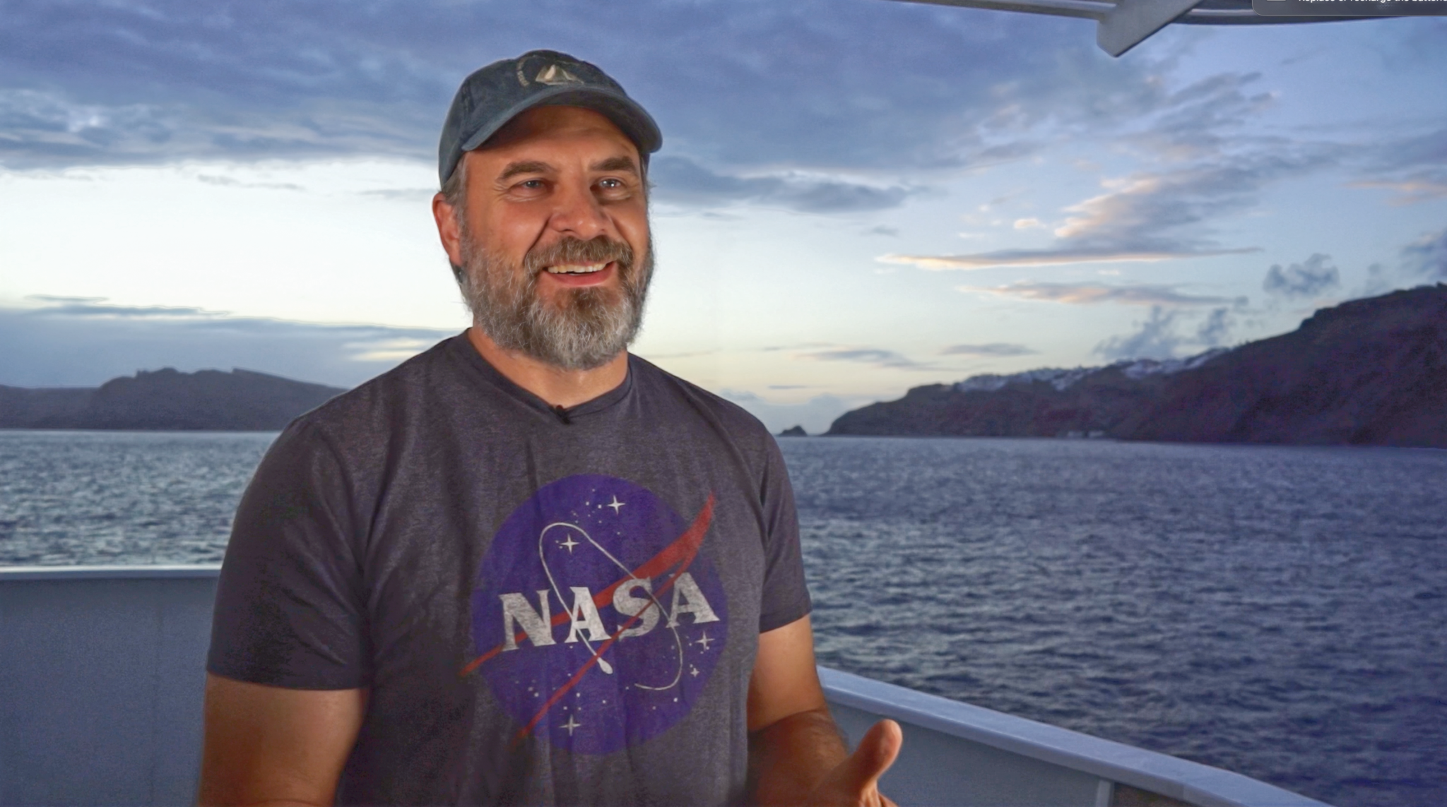 A middle-aged white man with a grayish-blonde beard smiles and stands on the deck of a ship, sun setting behind him. He is wearing a hat with a sailboat on it, and a gray NASA shirt.