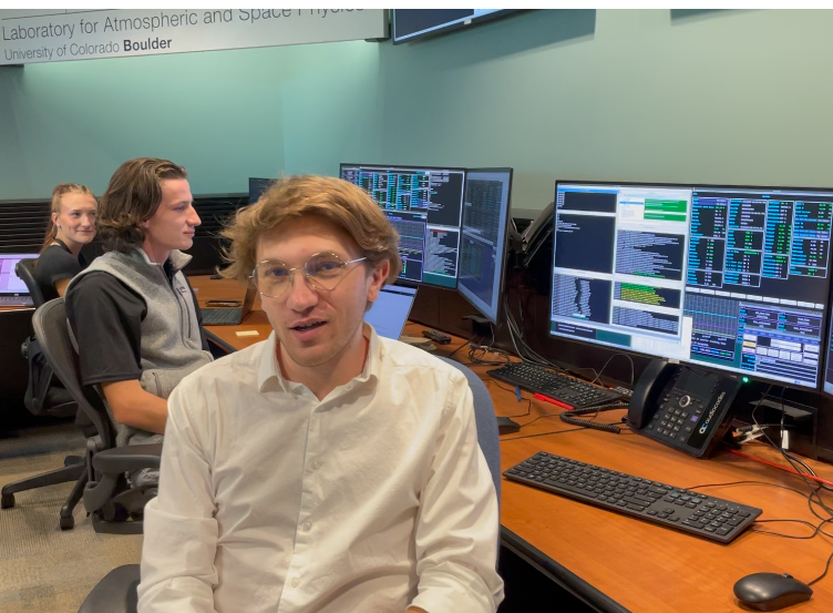 A young man with blonde hair and glasses and a white shirt sits in a control room. He is Sam Lippincott.