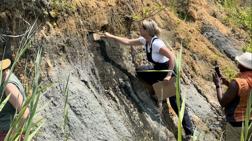 A group of people engaged in a fieldwork activity at a rocky outcrop. One person is using a brush and other tools to scrape the rock surface, while another person is recording the activity with a mobile phone. The setting is outdoors with tall grass and a layered rock formation in the background.