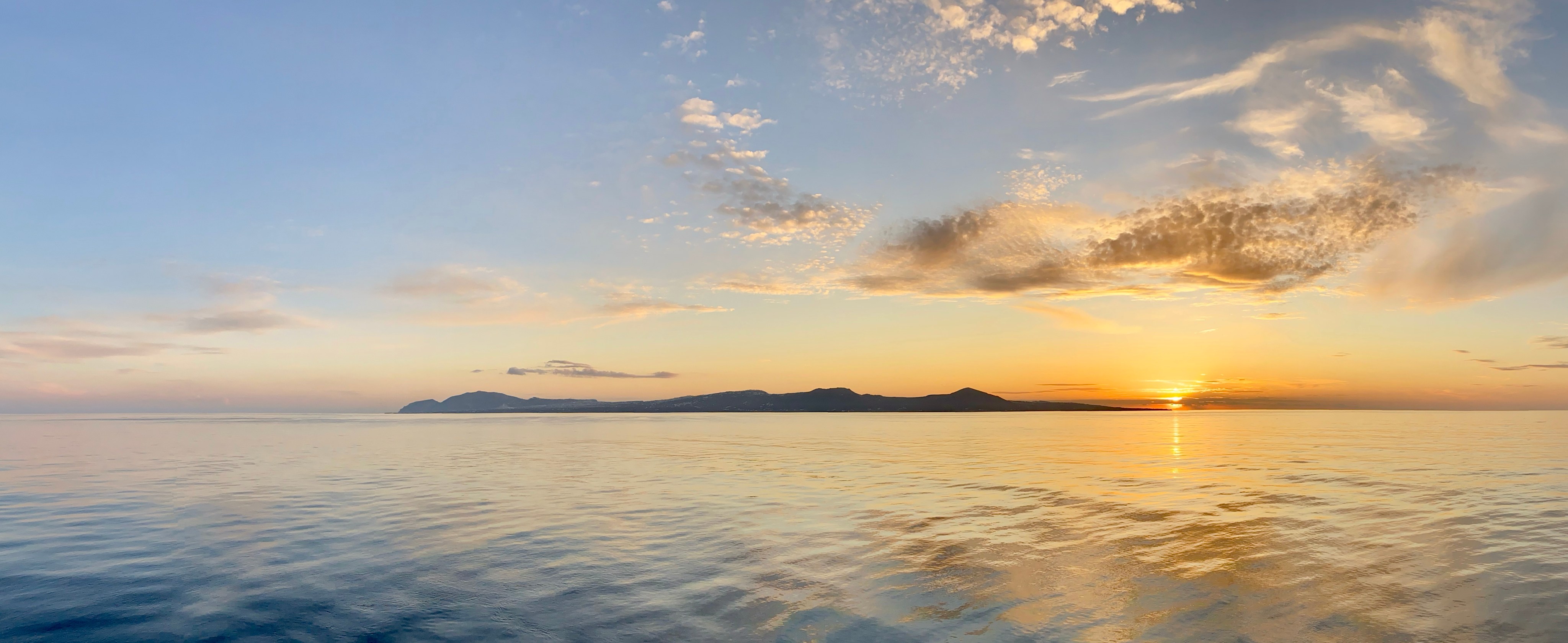 An ultrawide panorama of a sunrise at sea. The foreground shows a still ocean with minimal waves, receding to a small island on the horizon, with bright yellow and orange clouds against a blue sky.