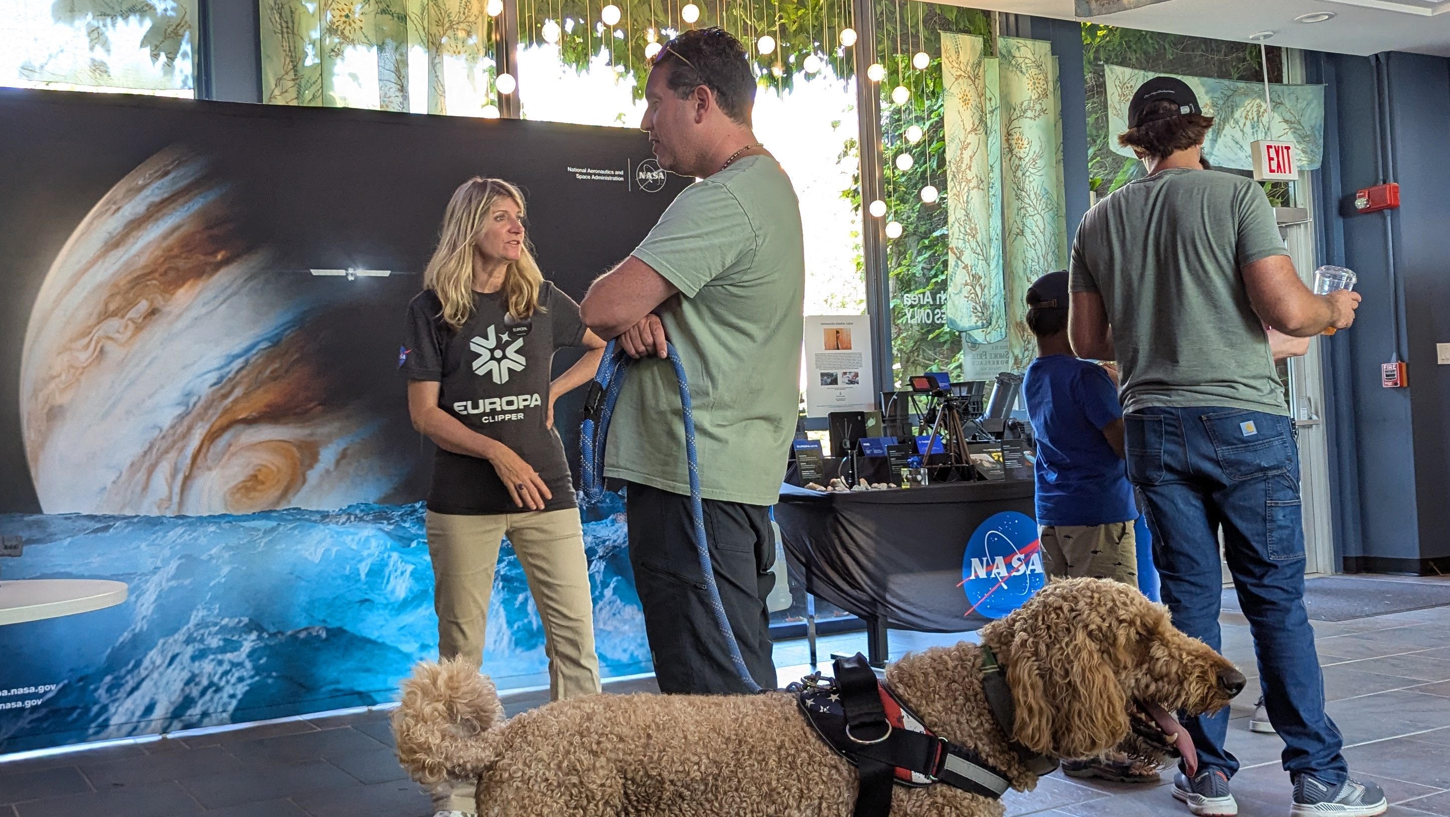 A woman wearing a Europa Clipper t-shirt talks to a man in a green shirt holding a leash to his dog at a NASA exhibit for the Europa Clipper mission. A man and boy stand nearby with their backs to the camera.