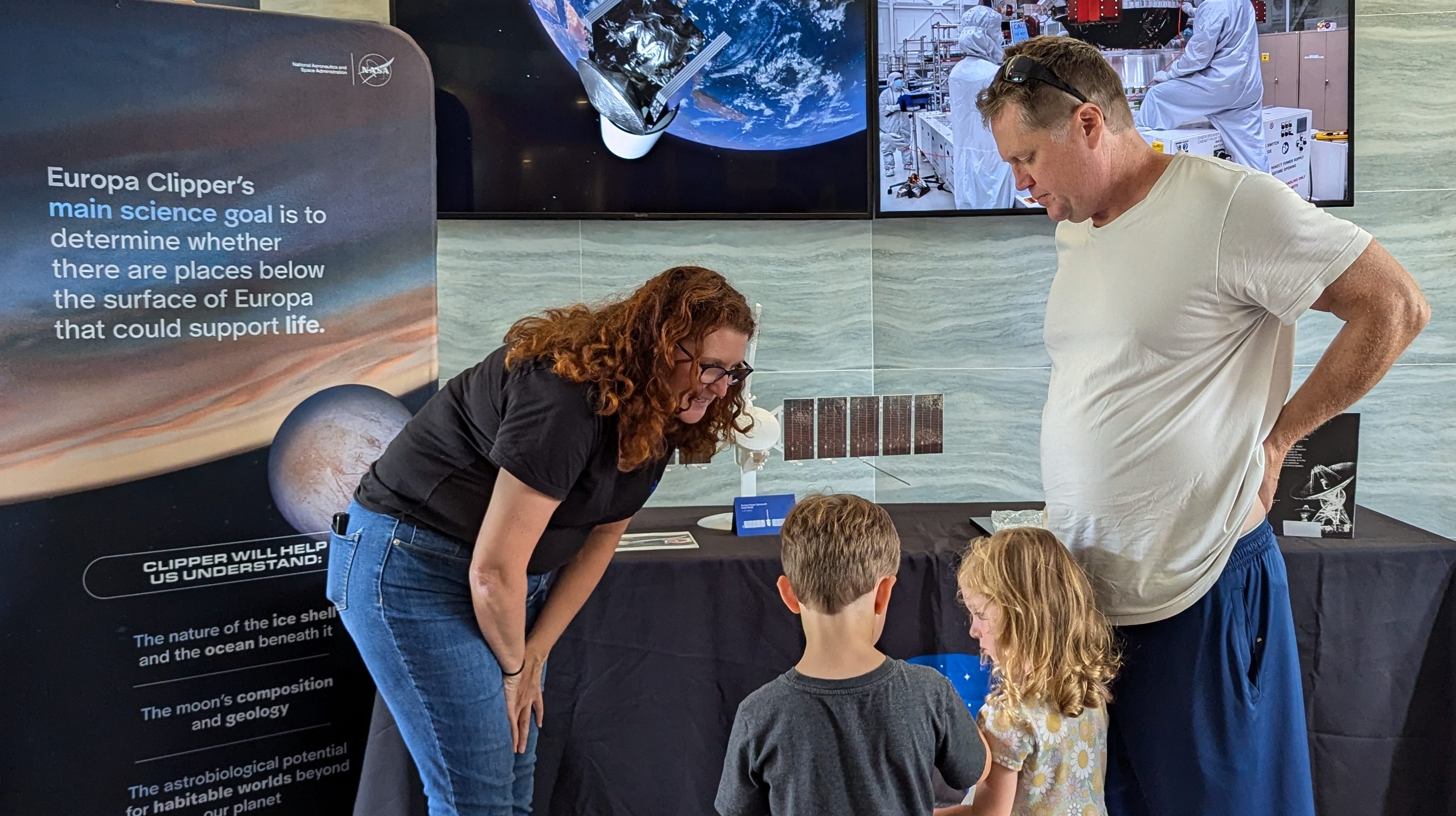 A NASA/JPL employee bends down to talk to two young visitors to NASA's Europa Clipper Roadshow as a man looks on.