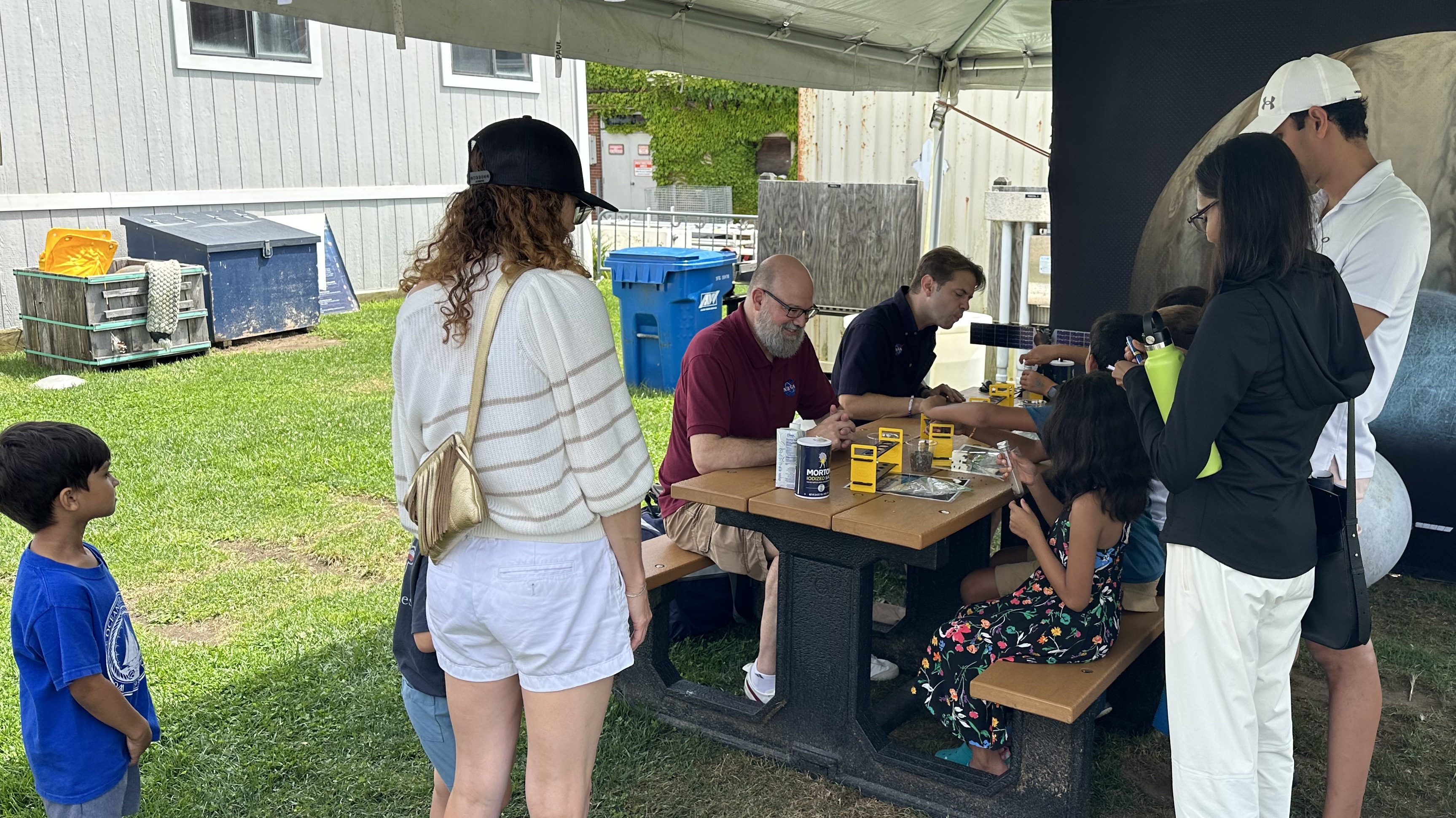 People sit at a picnic table to view experiments and talk to staff and volunteers. Other visitors stand nearby.