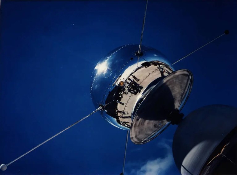 The Vanguard 1 satellite, a shiny, spherical object with protruding antennas, reflecting a group of people and the surrounding area under a clear blue sky.