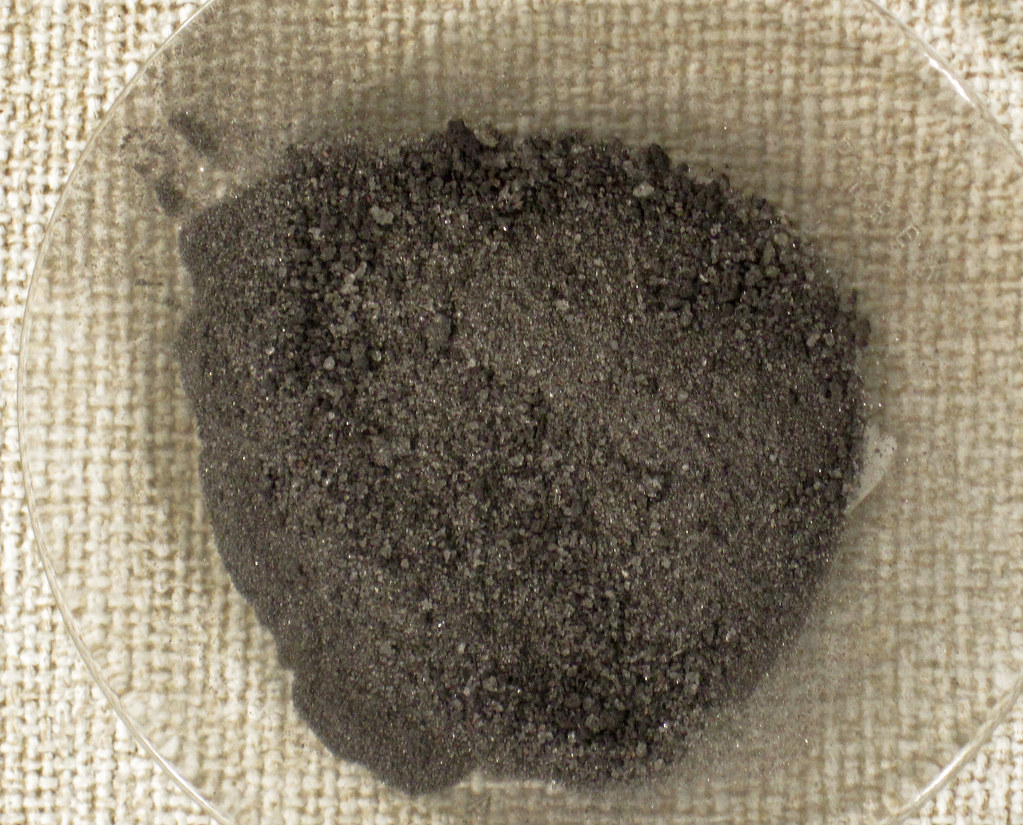 A close-up view of a small pile of dark gray powdered substance in a transparent shallow dish, placed on a beige textured surface. The grains of the powder vary in size, creating a rough and uneven appearance.