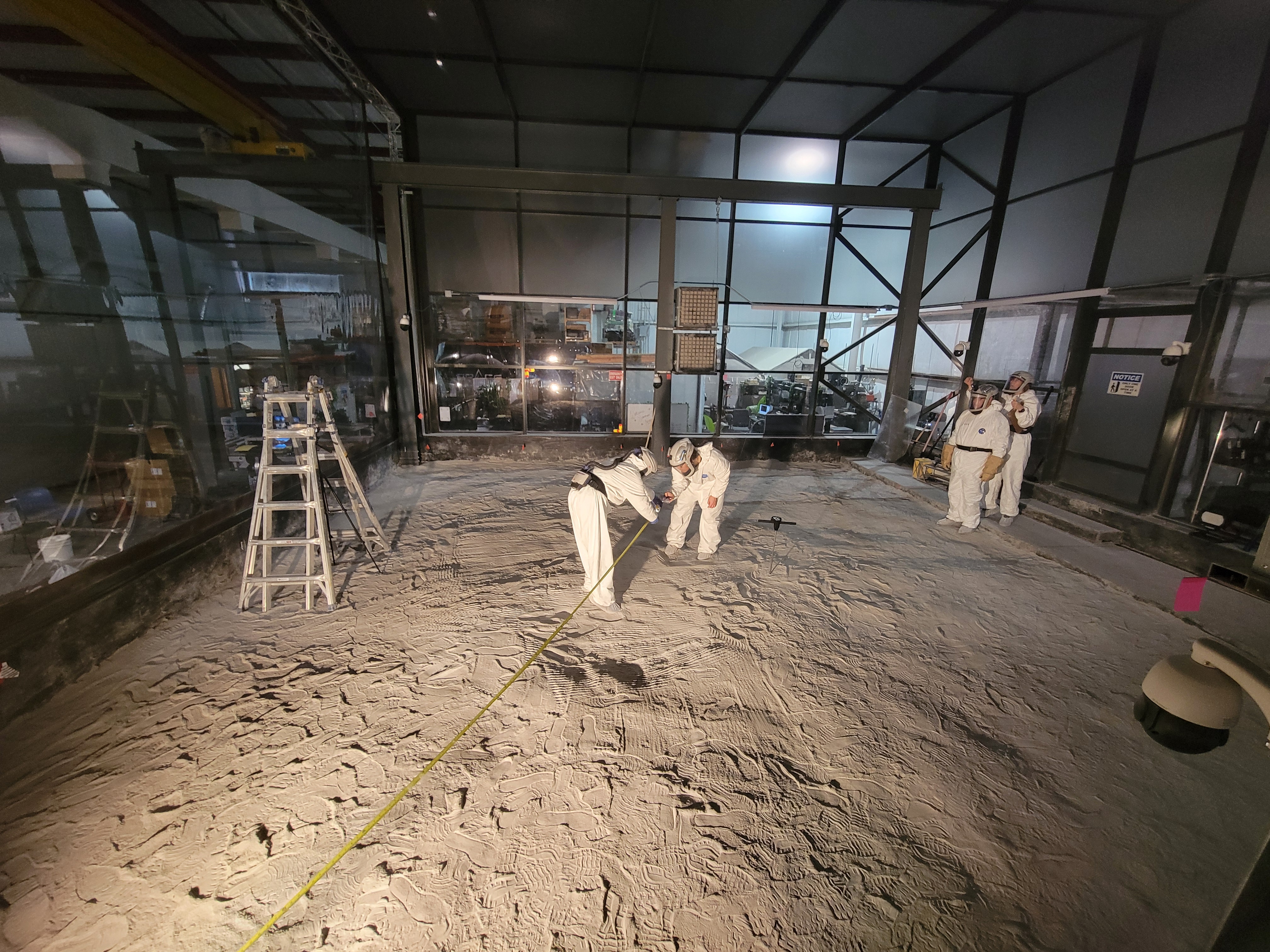 Several individuals wearing white, full-body suits with helmets are standing in what appears to be an industrial or laboratory setting. The ground is covered with a fine layer of sand or dust, which is marked with footprints and patterns from equipment. In the center of the image, two people are bending down and appear to be measuring something on the ground with a long yellow measuring tape. Their attention is focused on the sand or dust beneath them. To the right, in the background, two other individuals stand near the back wall, observing or possibly taking notes. A large glass wall on the left side reflects some of the lights in the room, which is otherwise dark. A silver ladder is standing on the left side of the room. The back of the room has industrial-style equipment, visible through the glass and within the setting. A light source is coming in sideways from outside the image, casting long shadows of the individuals and the objects in the room. The walls and ceiling are made of a metallic or industrial material, contributing to the laboratory or simulation vibe.