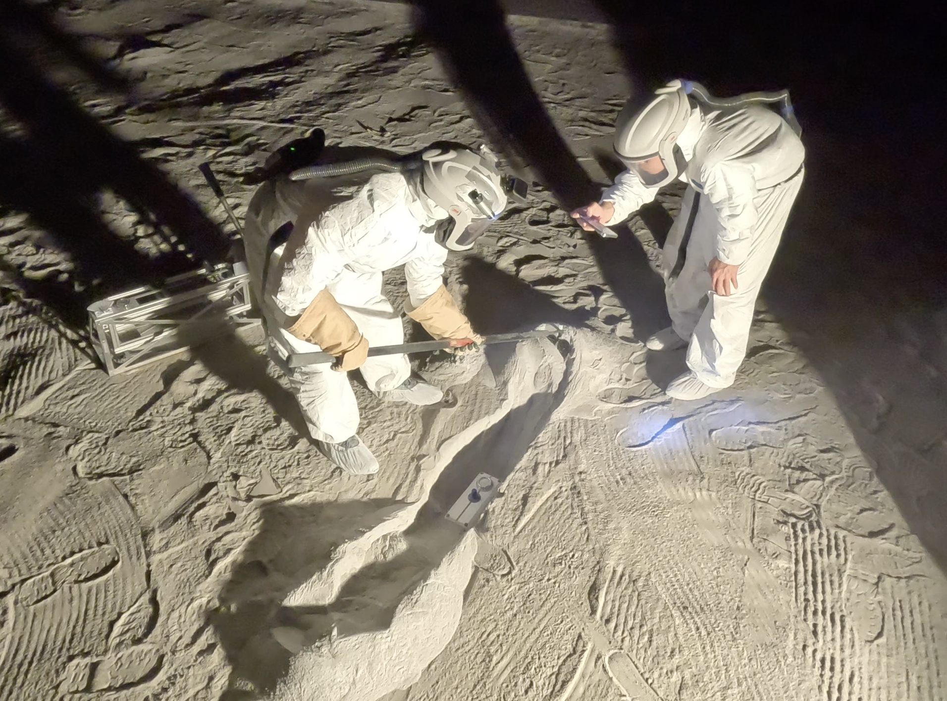 Two people dressed in bulky white suits and large helmets are standing on a sandy surface. One person is bending over and using a long tool to dig a shallow trench in the sand. The other person is also bending over the trench holding a flashlight and closely observing the area. The scene is dark, but the people are illuminated by an unseen light source, casting long shadows on the surface. The ground is textured with footprints and tool marks.