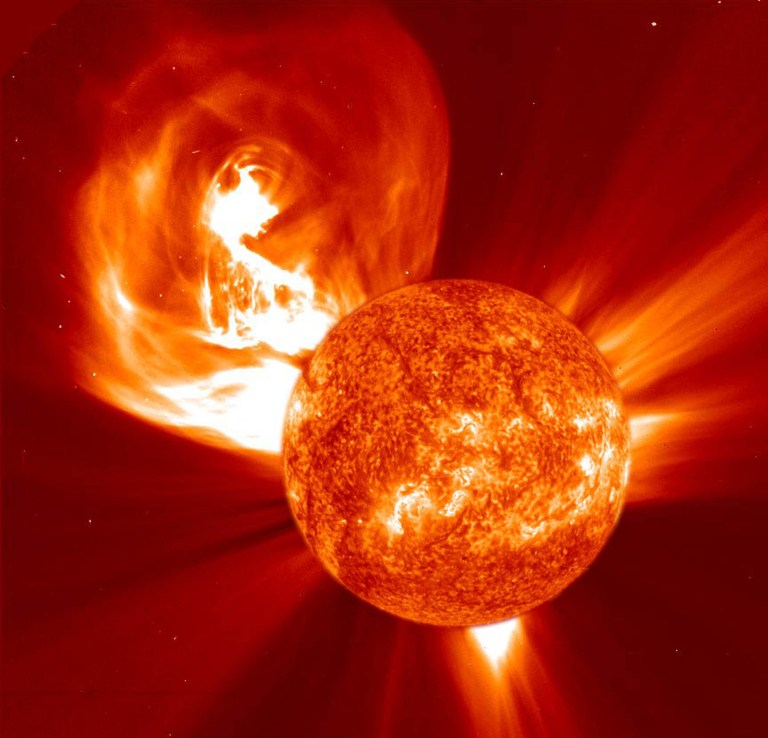 In the lower right, the Sun appears in red with blotchy, brighter areas of orange and yellow. In the upper left, a bulb-shaped cloud of material as large as the Sun extends away from the Sun. The inner parts of the cloud are bright white with orange and red streams of material around the edges.