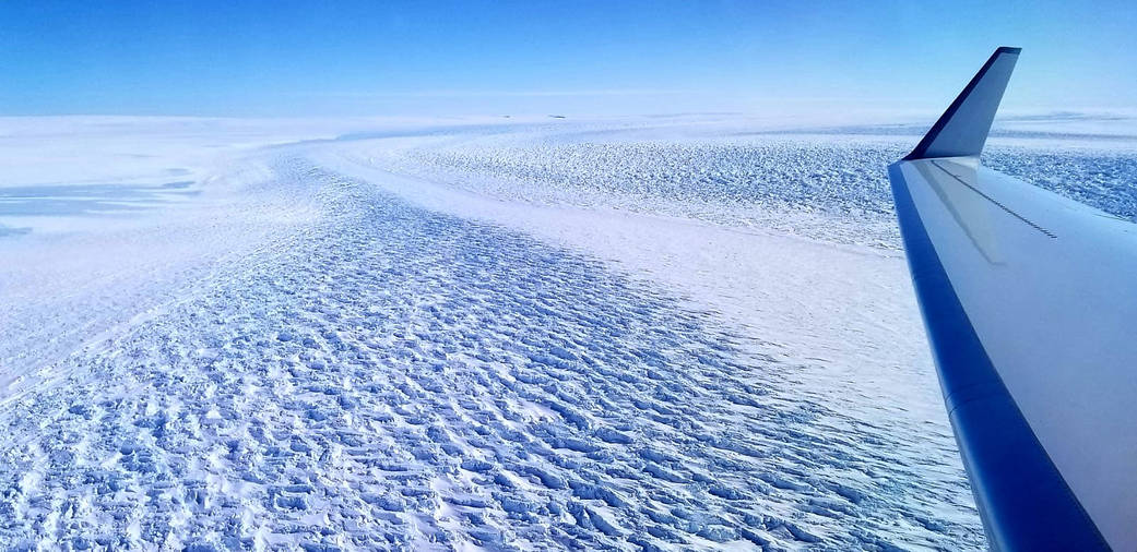 Aerial view of a vast, snow-covered glacier under a clear blue sky, with the wing of an aircraft visible on the right side of the image.