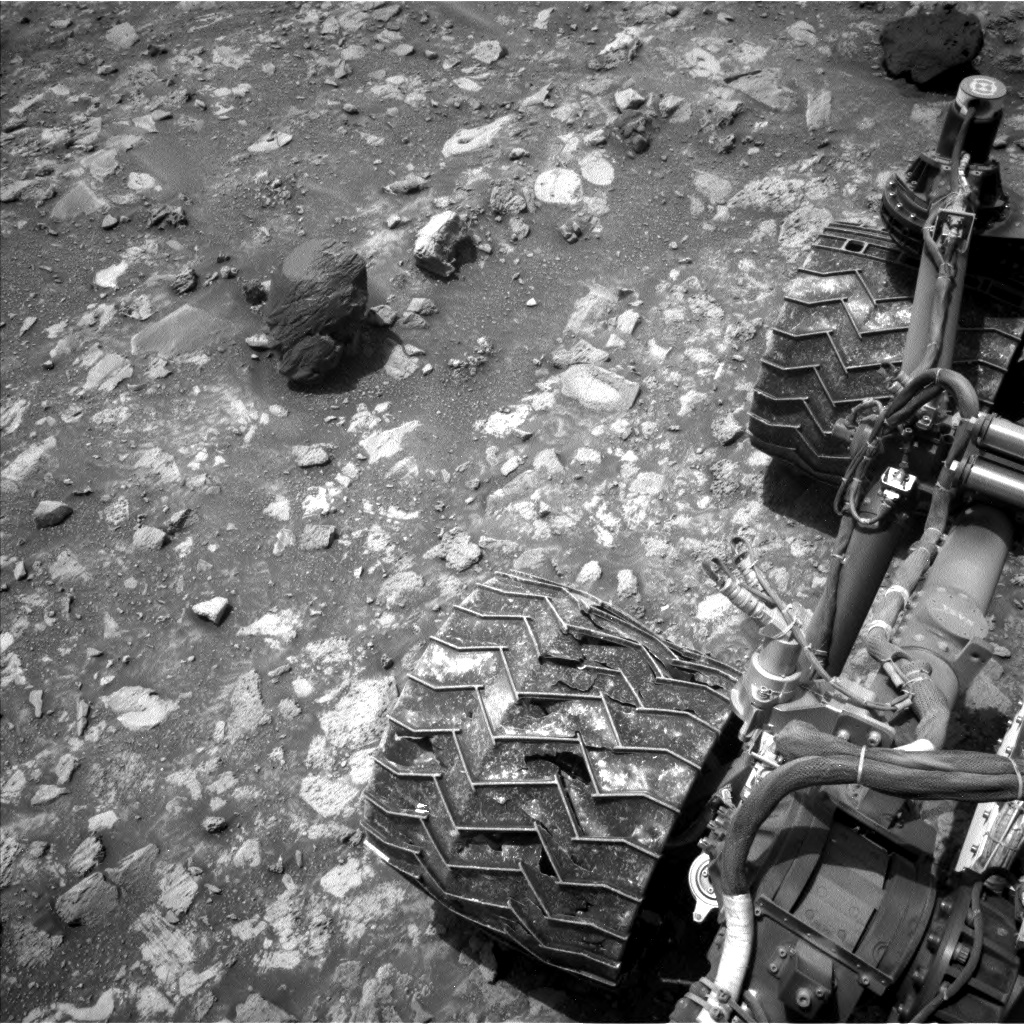 A grayscale photograph of the Martian surface shows medium gray soil interspersed with numerous light-colored rocks poking up from beneath the surface, on the left side of the frame, where one, much larger and darker rock, sits alone near the top of the frame. The right side of the image shows part of the rover, including two of its zig-zag treaded wheels, themselves covered in rocks and dust of various shades of gray.