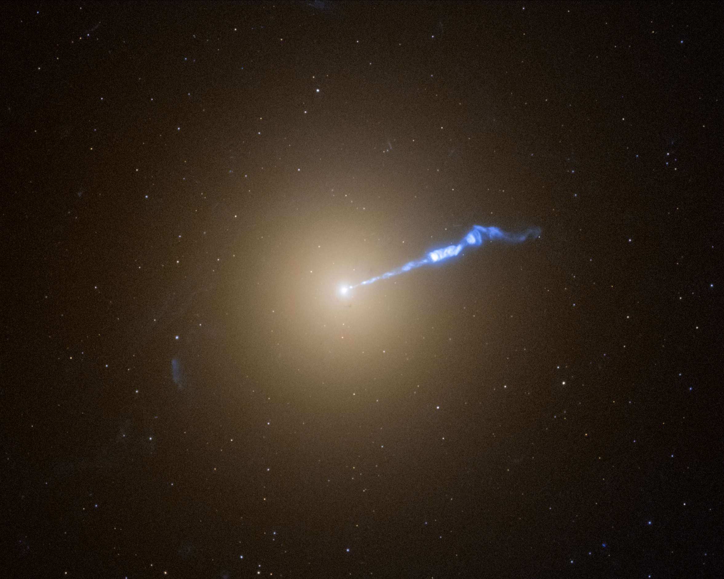 A Hubble image of galaxy M87, which resembles a translucent, fuzzy white cotton ball. The brightness decreases gradually out in all directions from a bright white point of light at the center. A wavy blue-white jet of material extends from the point-like core outward to the upper right, about halfway across the galaxy. Stars speckle the background.