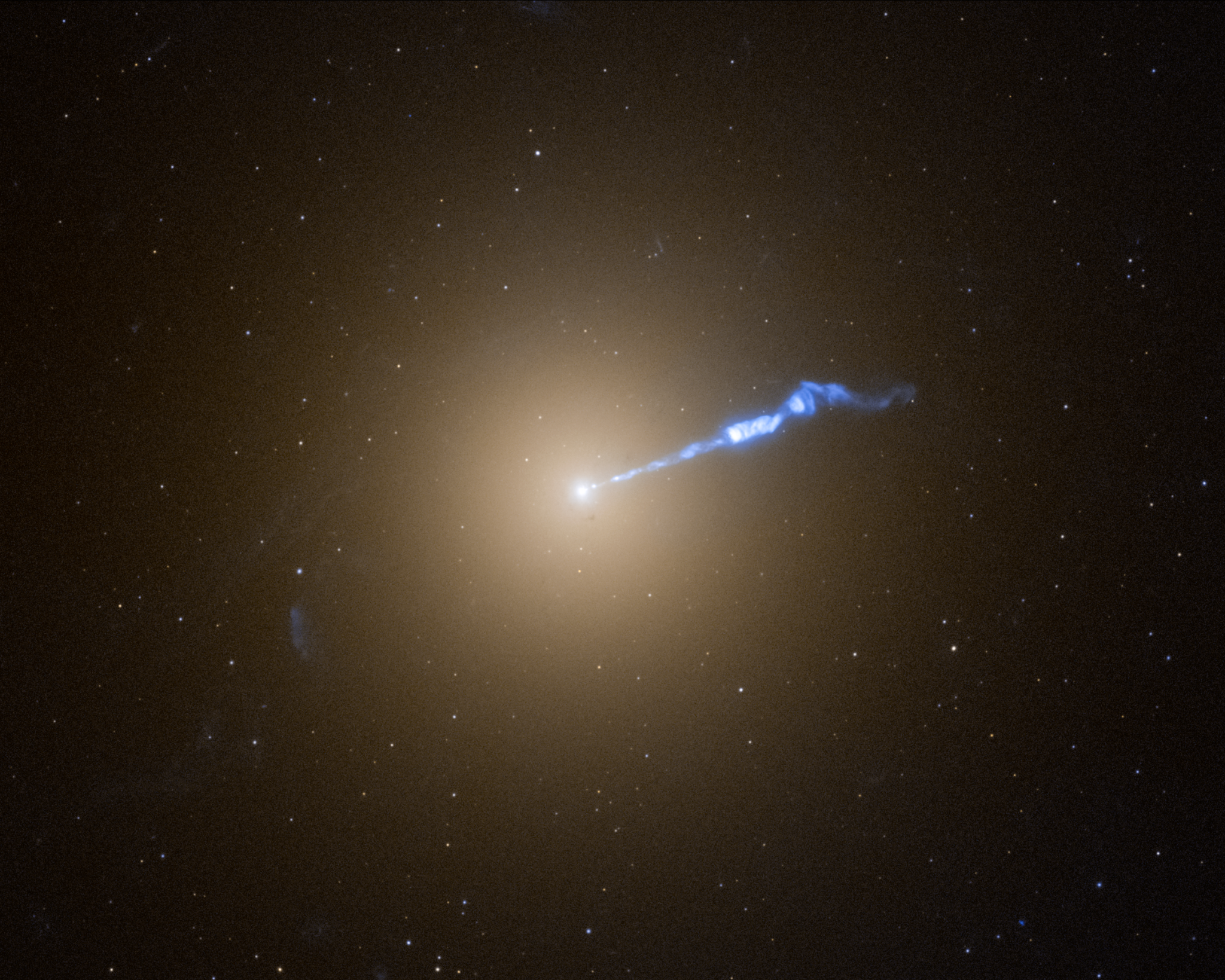 A Hubble image of galaxy M87, which resembles a translucent, fuzzy white cotton ball. The brightness decreases gradually out in all directions from a bright white point of light at the center. A wavy blue-white jet of material extends from the point-like core outward to the upper right, about halfway across the galaxy. Stars speckle the background.