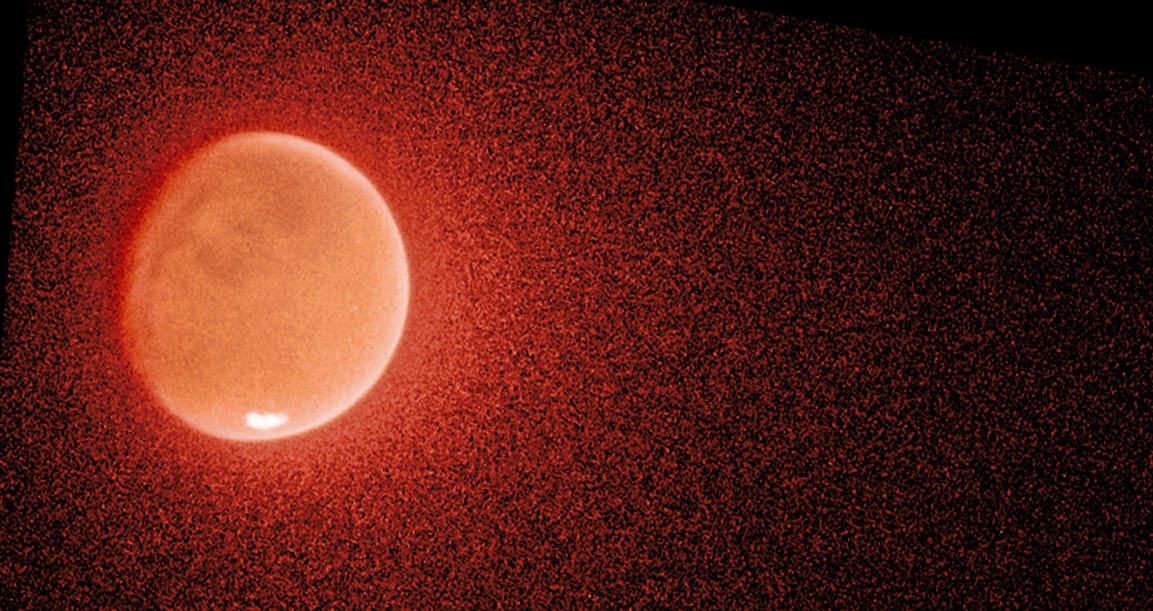 On a black background, a large orange and white orb is surrounded by a diffuse, grainy, orange halo. The halo appears to have more material on its right side than its left.