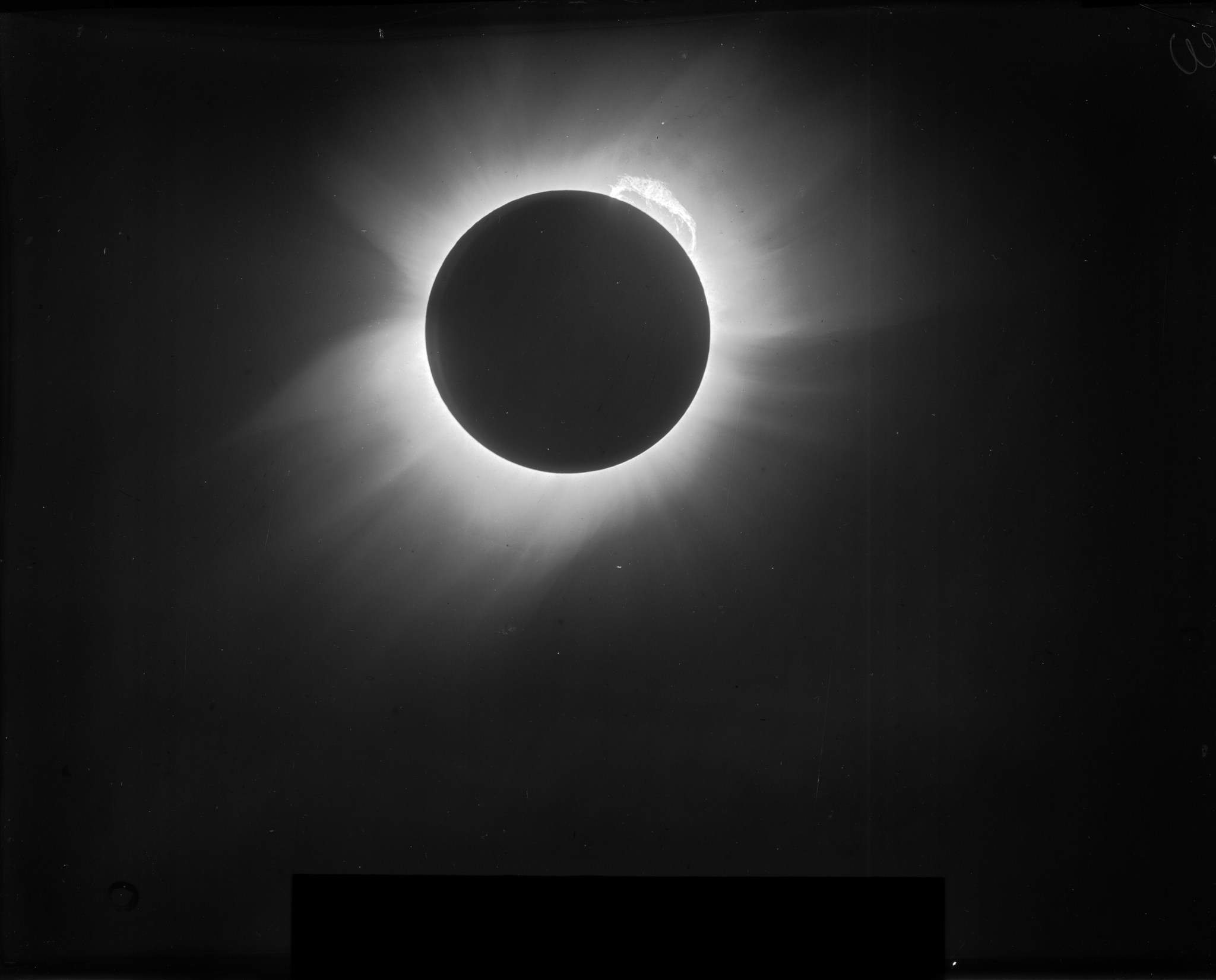 A black and white image of the total solar eclipse of May 29, 1919 taken from Sobral, Brazil. A large prominence arches across the upper right quadrant of the Sun's limb. Spikes of the Sun's outer atmosphere, the corona, ring its darkened disk.