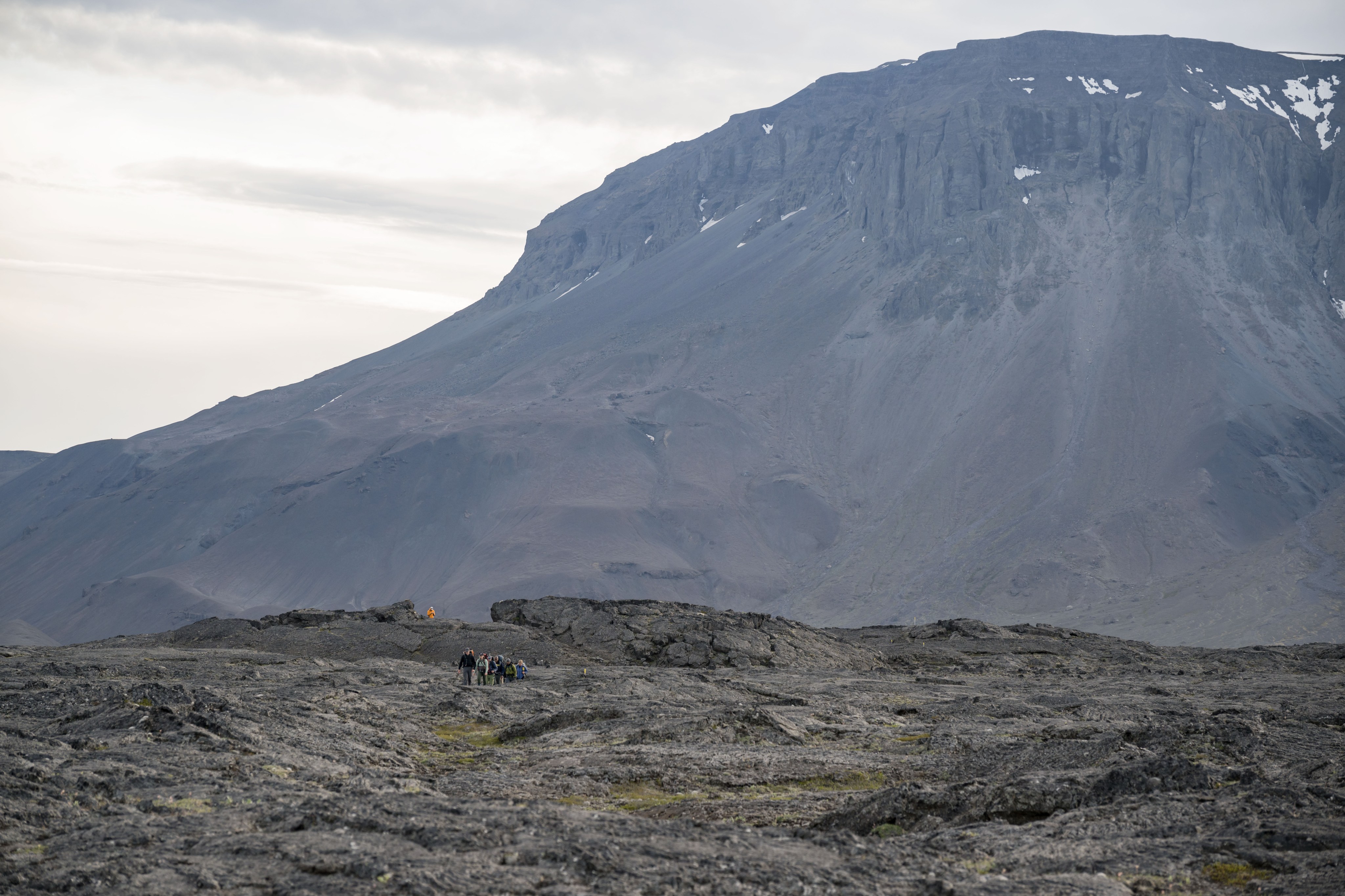 A group of hikers appears tiny against a sweeping basalt landscape. In the background, a steep, imposing hill topped with towering cliffs blocks out most of the sky.