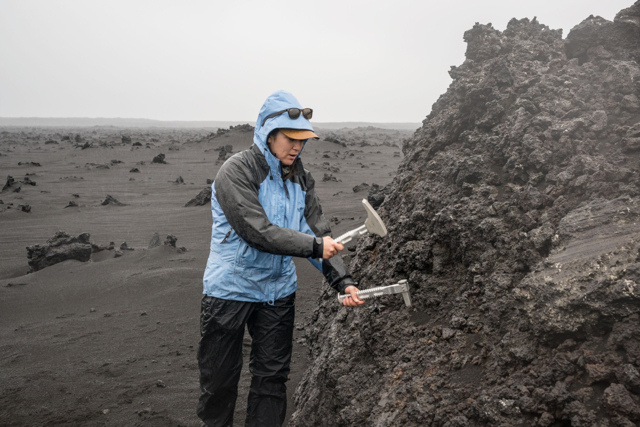 Researcher using hand-held tools to take a sample from a rugged basalt face. She wears outdoor work clothes and rain gear. In the background, dark grey dust and rocks stretch to the horizon. The landscape is unvegetated and entirely greyscale.