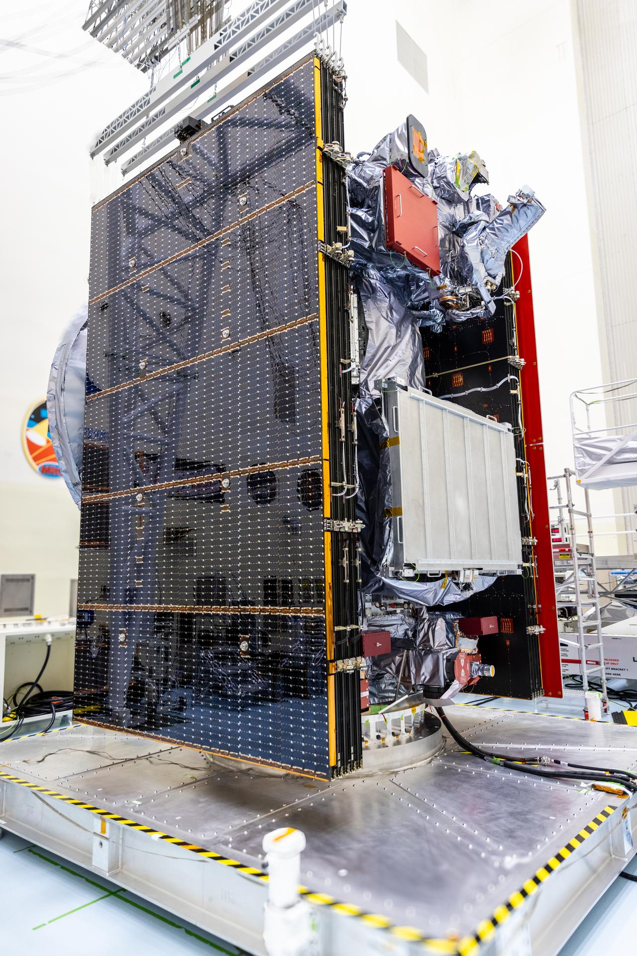 Europa Clipper sits on a silver work stand with yellow and black caution tape around the edges. One of the spacecraft's two solar wings is featured prominently in this image. It's folded up against the spacecraft body. The solar array has a shiny black surface with lots of yellow dots and stripes. It reflects part of the room.