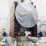 Europa Clipper sits on a silver work stand. The spacecraft's high gain antenna is in the front and center of the image. The folded solar panels are attached to each side of the spacecraft. At least four workers in blue jumpsuits and white head covers are to the sides of the spacecraft.