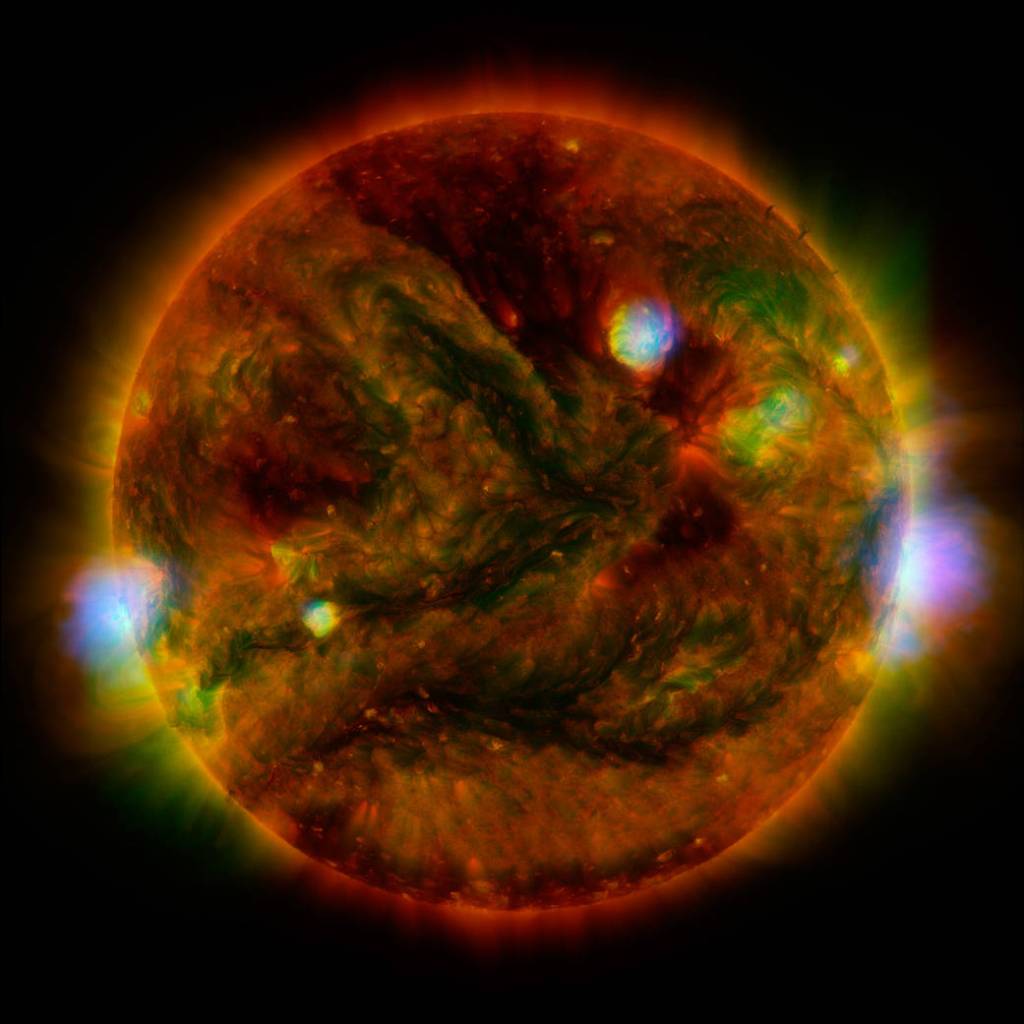 This image is a multi-wavelength composite of the Sun, showcasing its dynamic and turbulent surface. The Sun is depicted as a large, spherical object glowing with intense colors, including reds, greens, yellows, and bright blues. Bright, vibrant regions highlight areas of intense magnetic activity, while darker patches indicate cooler, less active areas. Wisps of solar material are visible swirling across the surface, creating a textured, almost swirling effect. The outer edges of the Sun appear to be radiating energy, with a glowing aura that extends into the surrounding blackness of space, illustrating the Sun's powerful solar flares and coronal emissions.