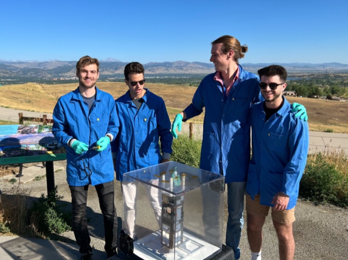 four team members wearing lab coats and protective gloves standing next to a rectangular structure housed in a clear cube.