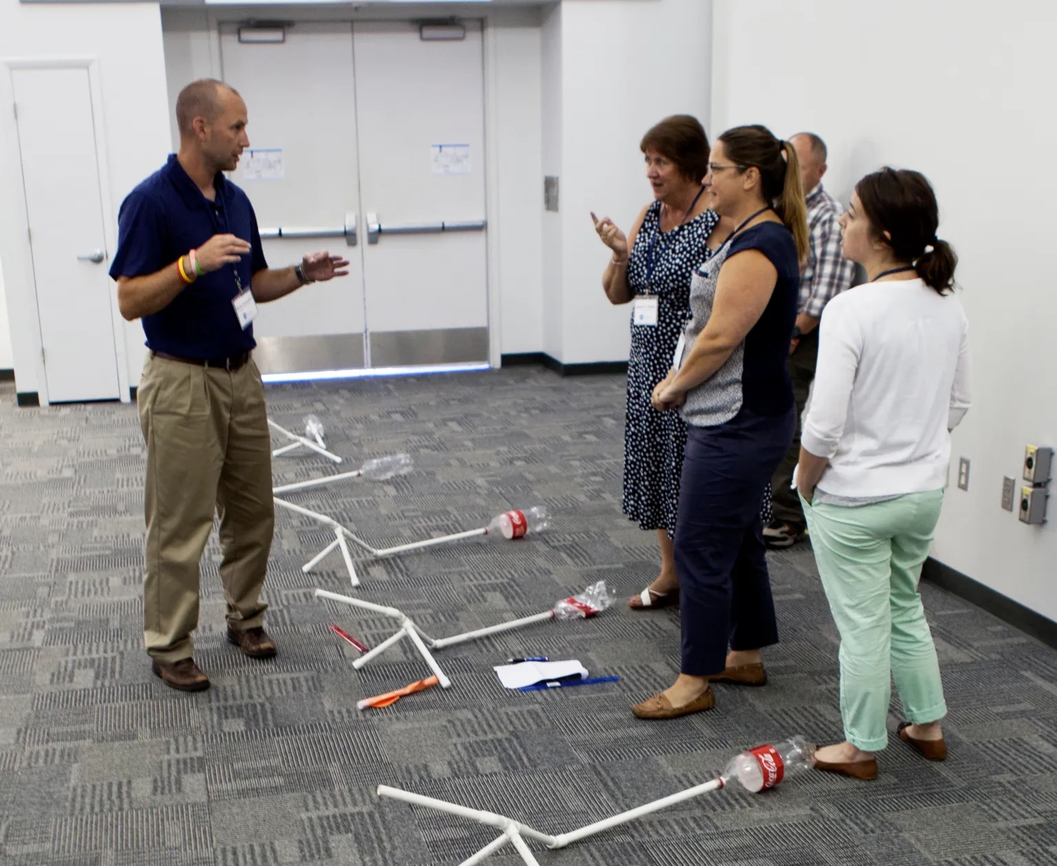 A group of four adults is standing in a room, listening to a man explaining something. The man is gesturing with his hands while speaking. The group is standing next to several white PVC pipe structures with empty soda bottles attached to the ends, which are scattered on the floor. The room has a carpeted floor and white walls with closed double doors in the background. The scene appears to be part of a workshop or training session.