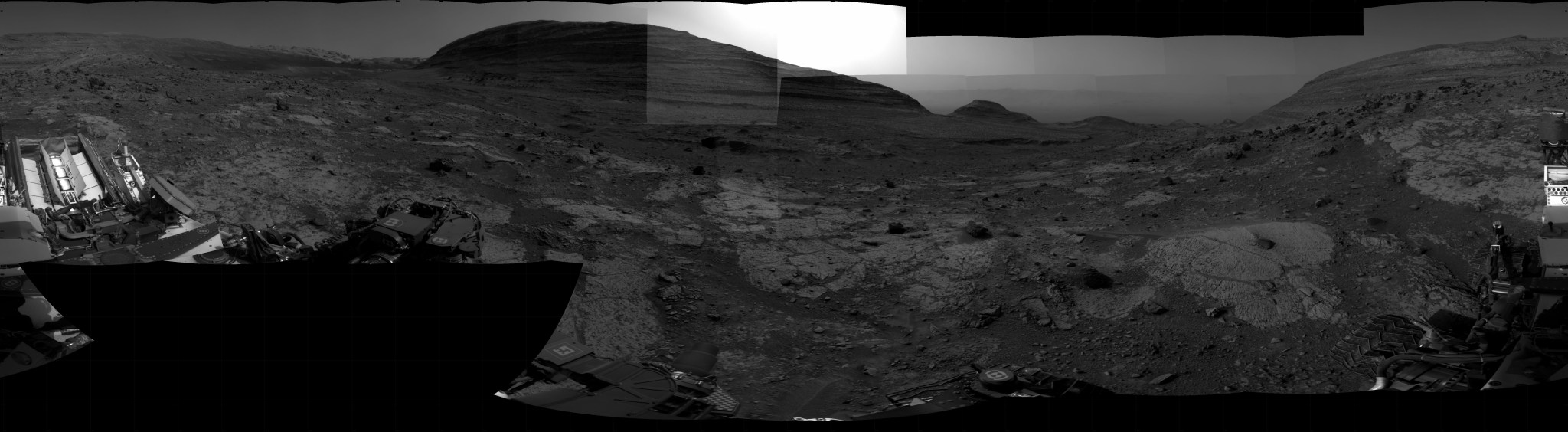 A grayscale panoramic image of the Martian surface shows darkening skies and ground, where the terrain is marked by uneven slabs of rock covered in some areas by soil and small rocks. A gently sloped, layered rock hill dominates the background in the top center of the image, hiding the Sun, which is illuminating the only bright area in the picture, the sky above and to the sides of the hill. Portions of the Curiosity rover are visible at the left, right, and bottom edges of the extreme wide-angle image.