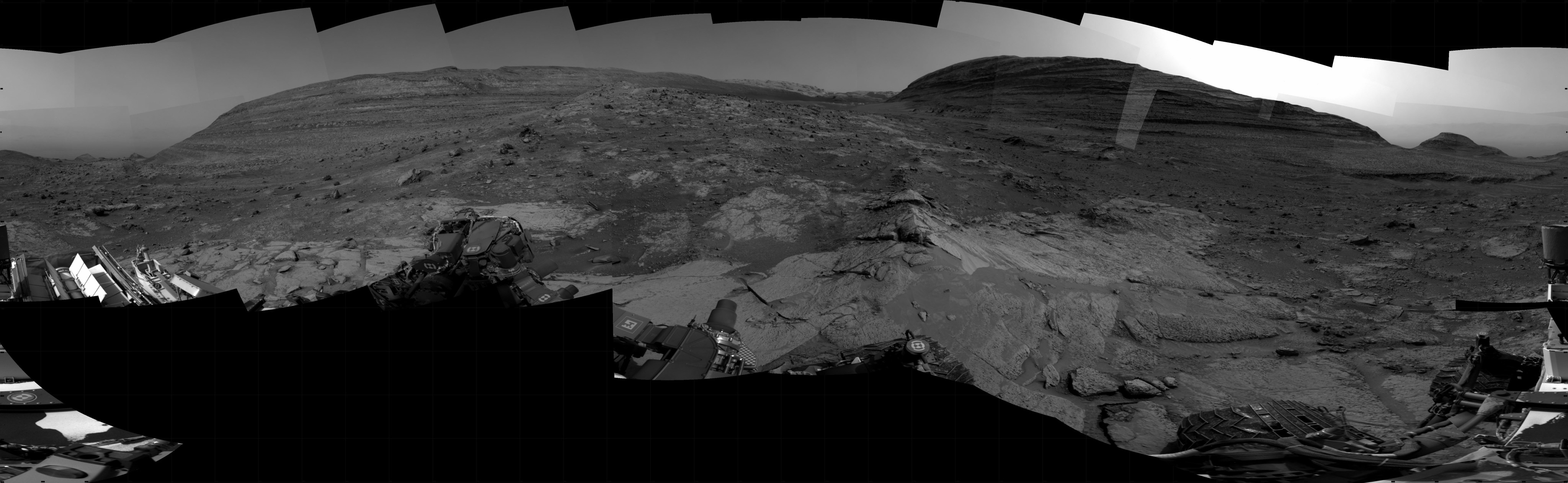 A grayscale panoramic image of the Martian surface shows darkening skies and ground, where the rolling terrain is marked by uneven slabs of rock covered in some areas by soil and small rocks. In the background, a gently sloped, layered rock hill rises above the ground at top right. To the left is a larger hill rising from the ground. The hill on the right is hiding the Sun, which is illuminating the only bright area in the picture, the sky above and to the sides of the hill. Portions of the Curiosity rover are visible at the left, right, and bottom edges of the extreme wide-angle image.