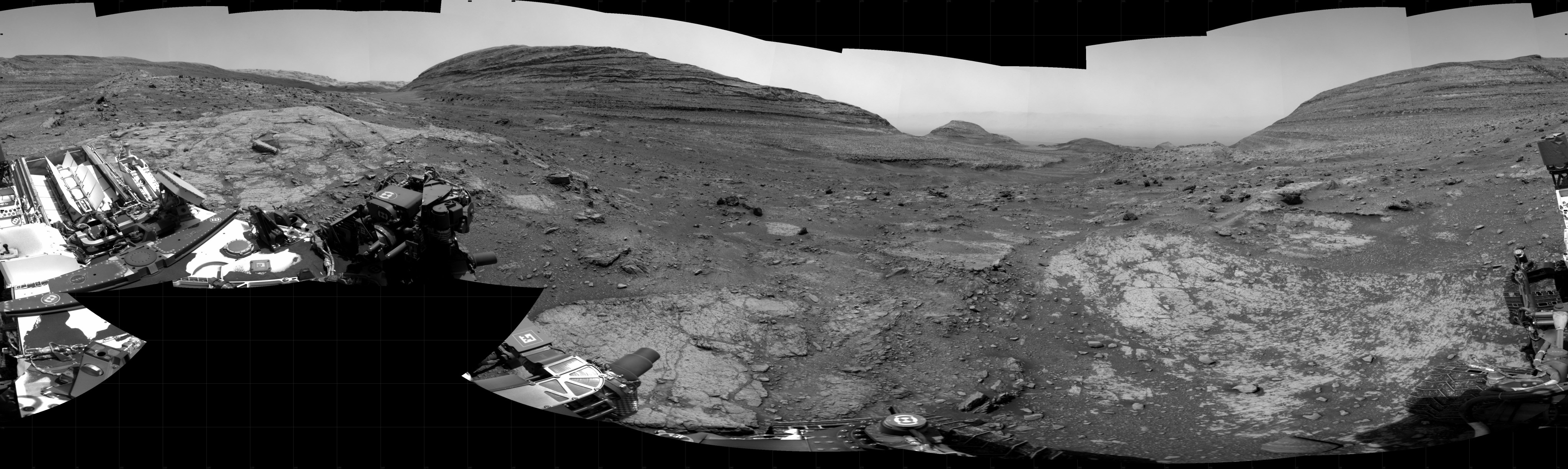 A grayscale panoramic image of the Martian surface shows booth sky and ground well-lit. The rolling terrain is marked by uneven slabs of rock covered in some areas by soil and small rocks. In the background, a wide, gently sloping, layered rock hill rises above the ground at center. Farther away, to the right, is another hill with a more pointed top, and at third rises up to the right edge of the frame. Portions of the Curiosity rover are visible in the frame of this extreme wide-angle image, taking up most of the lower-left corner. Several instruments and pieces of equipment are visible atop the rover, as are logos for NASA and JPL, with very dark, fine dust covering some areas.