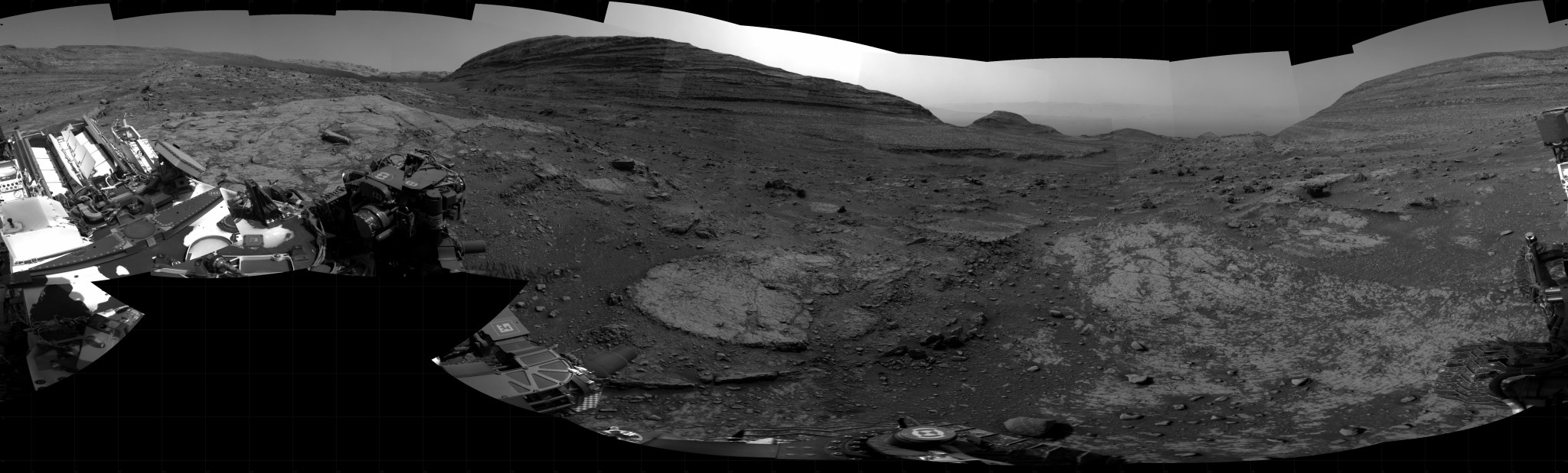 A grayscale panoramic image of the Martian surface shows booth sky and ground well-lit. The rolling terrain is marked by uneven slabs of rock covered in some areas by soil and small rocks. In the background, a wide, gently sloping, layered rock hill rises above the ground at center. Farther away, to the right, is another hill with a more pointed top, and at third rises up to the right edge of the frame. Portions of the Curiosity rover are visible in the frame of this extreme wide-angle image, taking up most of the lower-left corner. Several instruments and pieces of equipment are visible atop the rover, as are logos for NASA and JPL, with very dark, fine dust covering some areas.