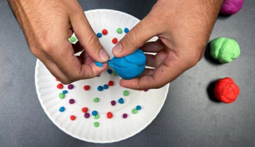 A set of hands tearing apart blue modeling clay over a paper plate with other pieces of modeling clay on it in purple, green, and red.