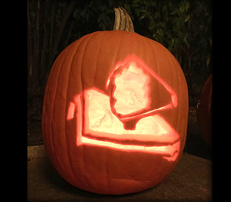 View of an illuminated jack-o'-lantern style pumpkin carved with an image of overview of the James Webb Space Telescope with its distinctive sunshield and mirror components.