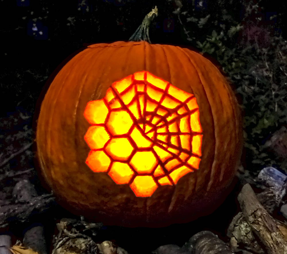 View of an illuminated jack-o'-lantern style pumpkin carved with an image of the hexagon mirror segments of the James Webb Space Telescope with a spider web overlaying the top right portion of the overall collection of mirror segments.