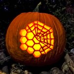 View of an illuminated jack-o'-lantern style pumpkin carved with an image of the hexagon mirror segments of the James Webb Space Telescope with a spider web overlaying the top right portion of the overall collection of mirror segments.