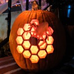 View of an illuminated jack-o'-lantern style pumpkin carved with an image of the 18 hexagon mirror segments of the James Webb Space Telescope with a spider figure in the top right portion of the overall collection of mirror segments.