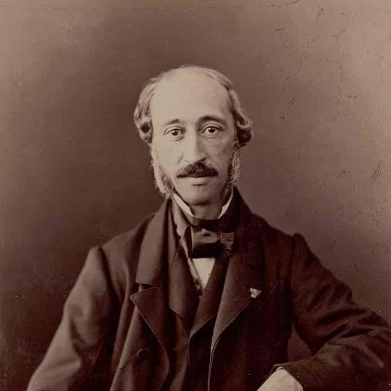 A sepia-toned vintage photograph of a middle-aged man with receding hair, prominent sideburns, and a mustache. He is dressed formally in a dark suit with a bow tie, sitting against a plain backdrop.