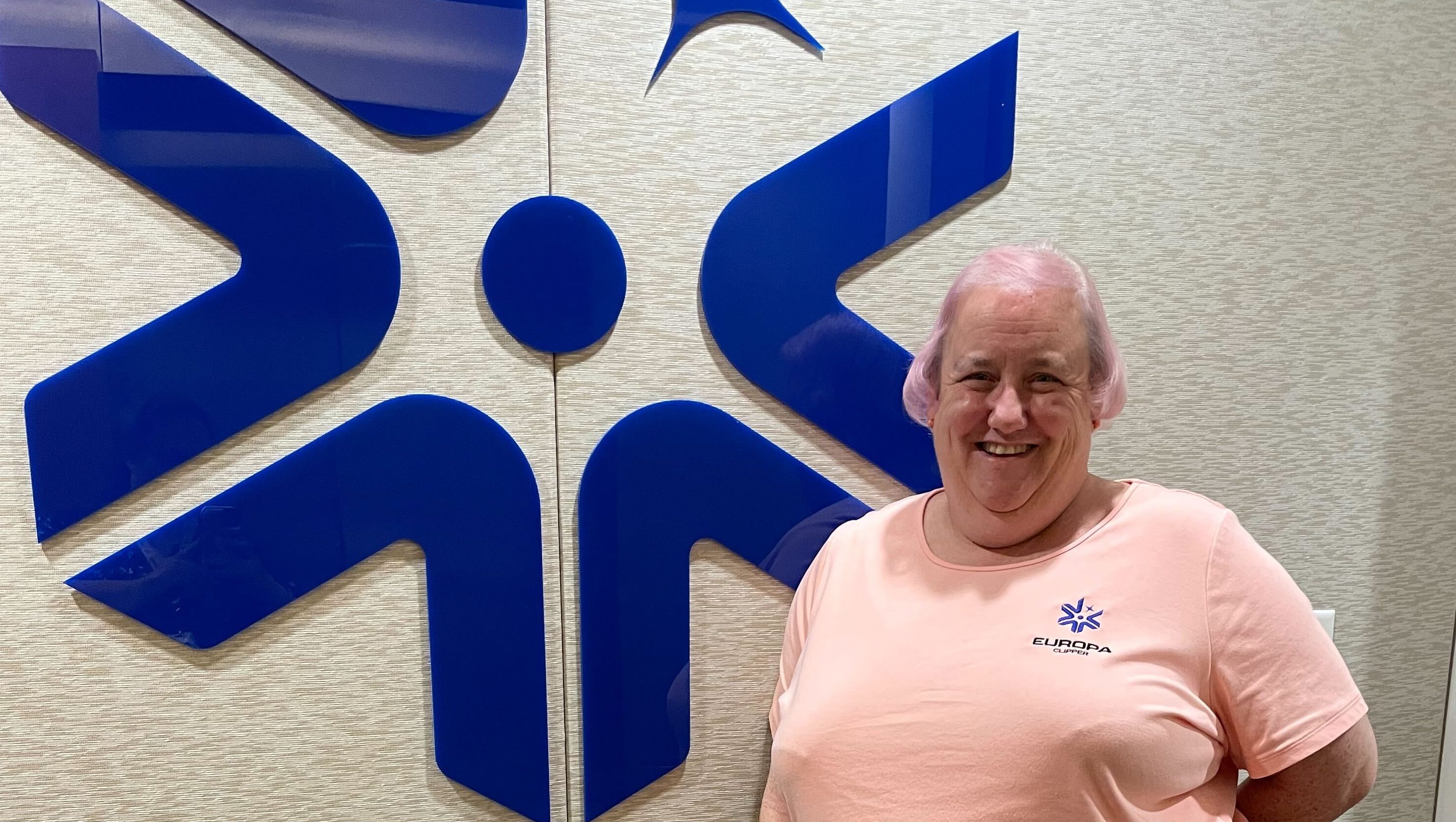 Trina Ray wearing a pink shirt with the blue Europa Clipper logo, and standing in front of a similar logo on a wall.