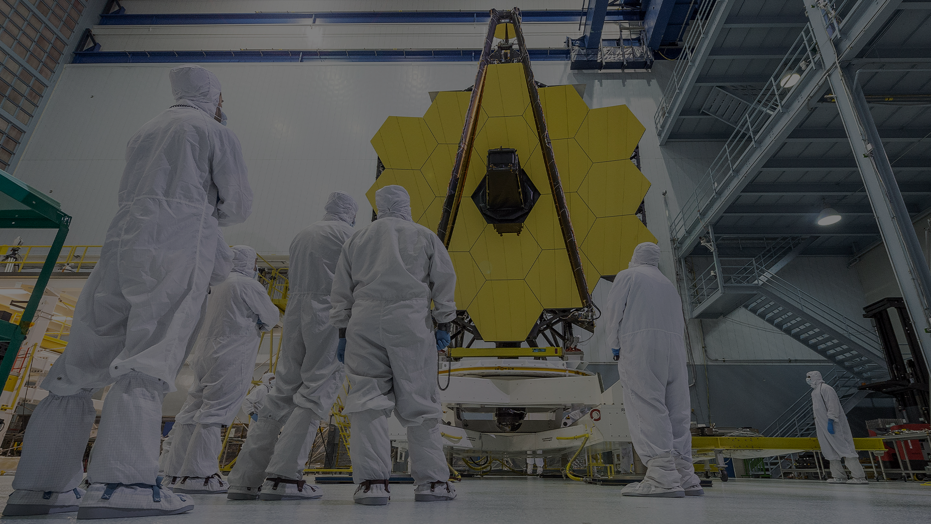 Ground-level darkened view of Webb Primary Mirror with technicians in foreground in the Goddard Clean Room.