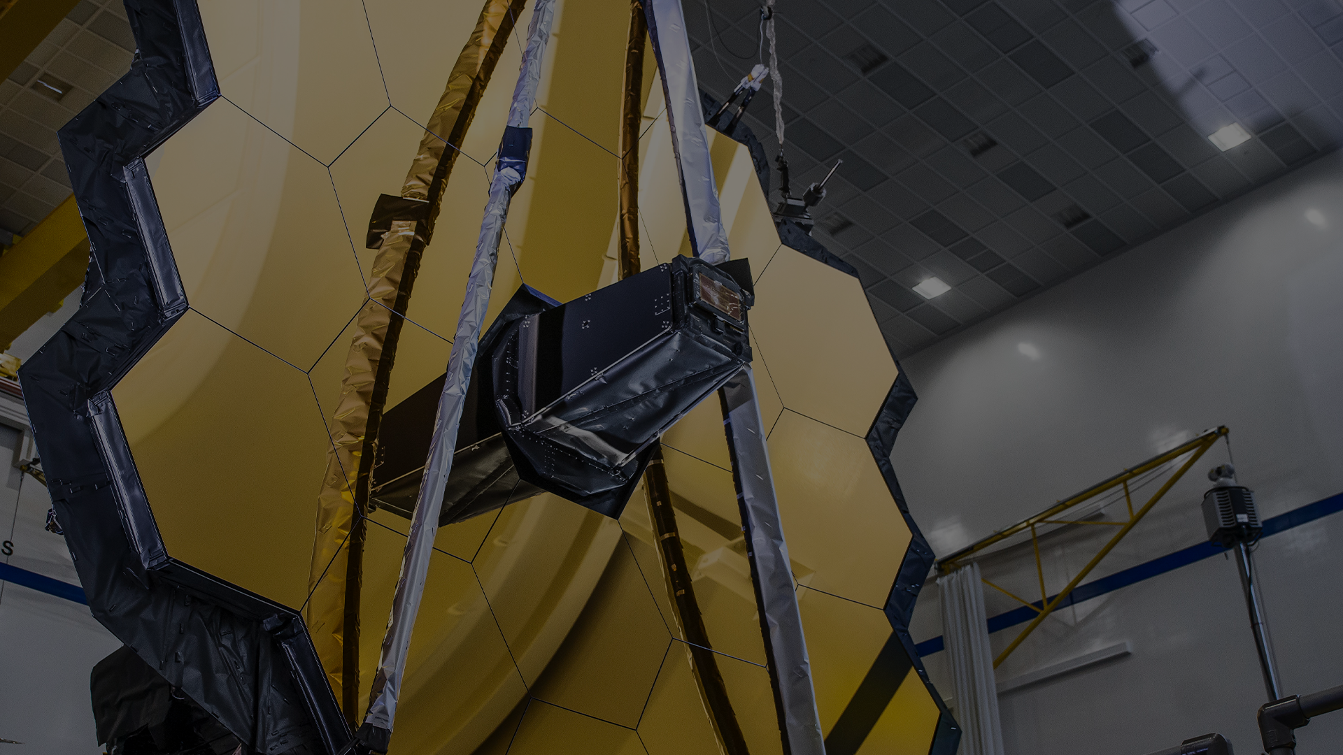 Super closeup darkened view of Webb Primary Mirror in the left side foreground and some clean room structures in the right side background.