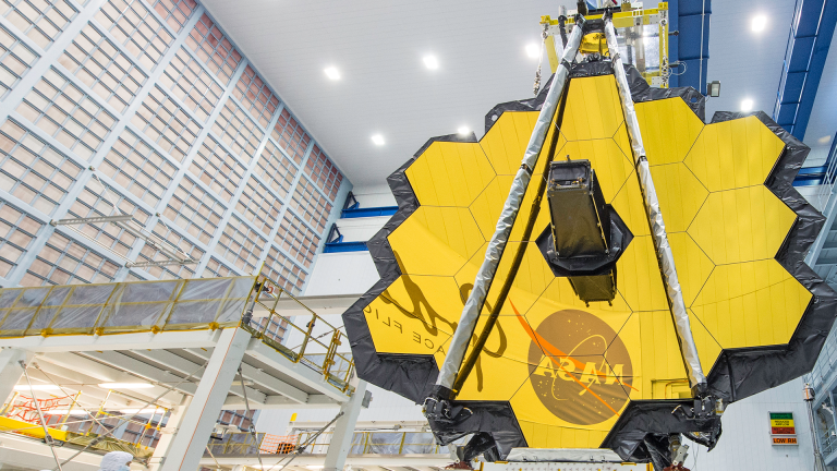 Overview of Webb Primary Mirror in right side foreground with the NASA meatball logo and part of the Goddard logo reflecting in the Webb mirror segments and with clean room structures shown in the left side background.