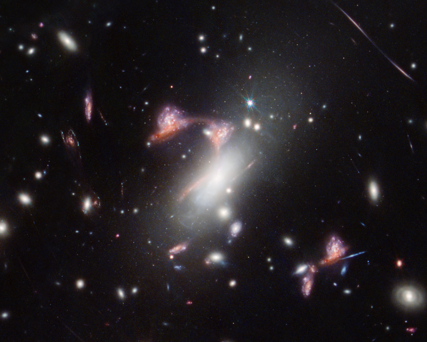 Amid a field of galaxies, a repeated, elongated red galaxy forms a shape like the top of a question mark, with another galaxy positioned like the question mark’s dot. In each occurrence, another white, clumpy galaxy with an overall circular shape appears perched on top of the red galaxy. A very bright foreground galaxy appears to the right of the bottom curve of the question mark shape. To the lower right, among other galaxies, another occurrence of the galaxy pair appears, unaffiliated with the question mark shape.