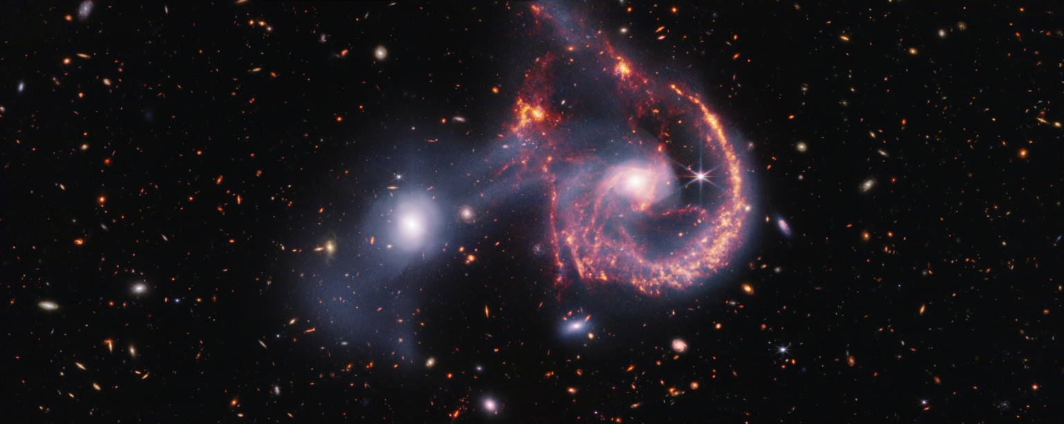 A pair of interacting galaxies. The larger of the two galaxies is slightly right of center, and composed of a hazy, bright, white center and a ring of gaseous filaments, which are different shades of red and orange. Toward the bottom left and bottom right of the ring are filaments of gas spiraling inward toward the core. At the top left of the ring is a noticeable gap, bordered by two large, orange pockets of dust and gas. The smaller galaxy to its left is made of hazy white gas and dust, which becomes more diffuse farther away from its center. To this galaxy’s bottom left, there is a smaller, more diffuse gas cloud that wafts outward toward the edges. Many red, orange, and white galaxies are spread throughout, with some hazier in composition and others having more defined spiral patterns.