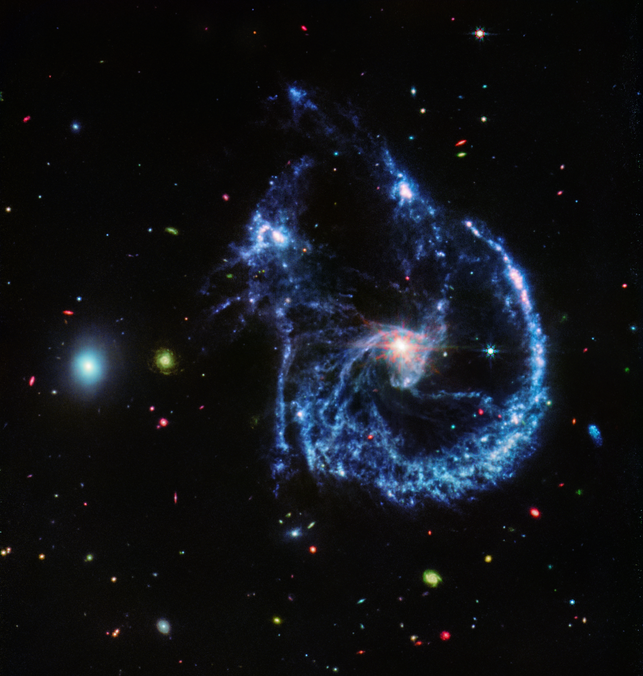 A pair of interacting galaxies. The larger of the two galaxies is slightly right of center, and is composed of a bright, white center and a ring of blue, gaseous filaments. The center of this galaxy shows Webb’s eight-pronged diffraction pattern. There are three filaments of gas and dust moving from the ring toward the center. At the top left of the ring is a noticeable gap, bordered by two large, blue pockets of dust and gas. The smaller galaxy is made of hazy, light blue gas and dust. Many red, green, blue, and yellow galaxies are spread throughout, with some being hazier in composition and others having more defined spiral patterns.