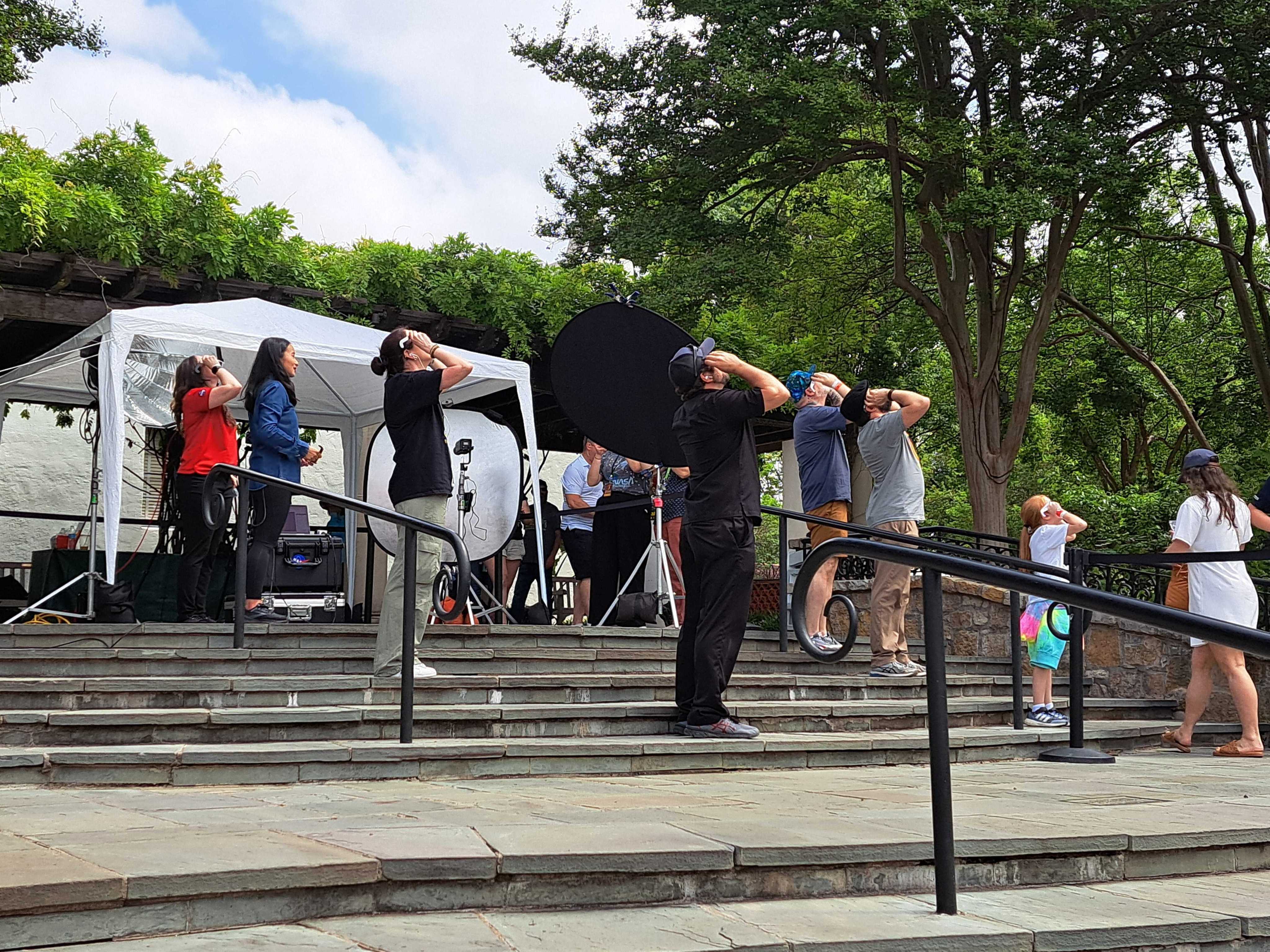 A group of people stand on outdoor stairs. They are looking up at the sky while wearing eclipse glasses. They are standing among video and lighting equipment.