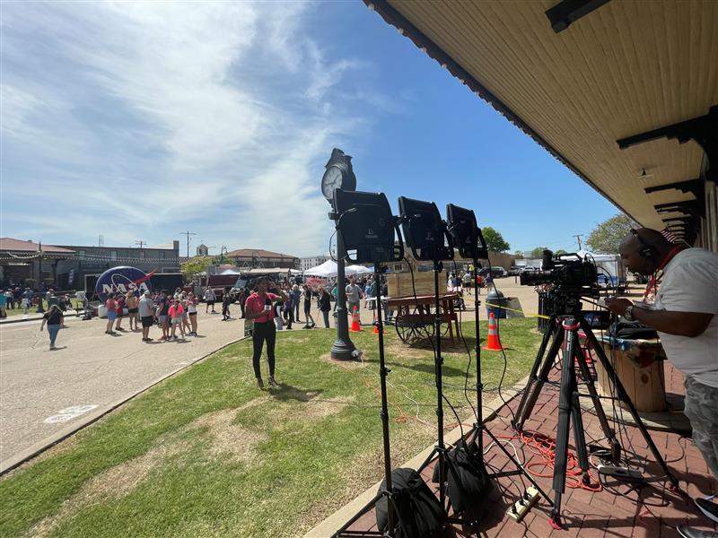 People tinker with camera equipment in front of a grassy and concrete area with people scattered about.