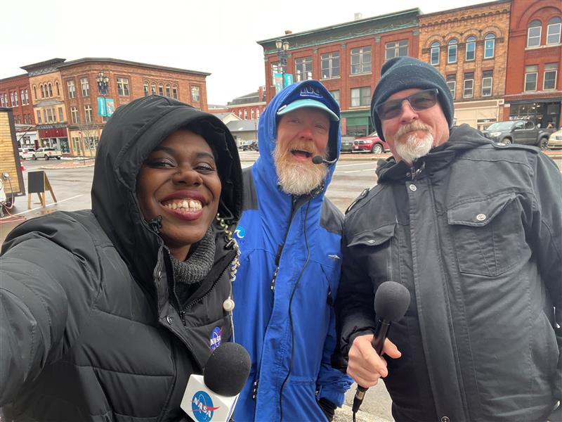 Three people smile for a selfie. One is holding a microphone. Behind them are brick buildings.