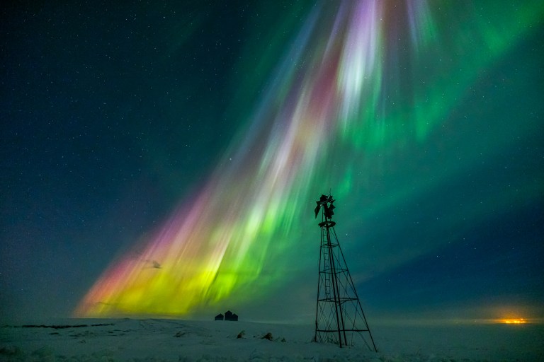 A dazzling display of multicolored auroras stretches diagonally across the sky, featuring vibrant green, yellow, pink, and purple hues. The lights cascade over a snowy landscape, with a silhouetted windmill and distant structures adding depth to the scene. The night sky is dotted with stars, enhancing the ethereal beauty of the auroras, which appear to radiate toward the horizon.