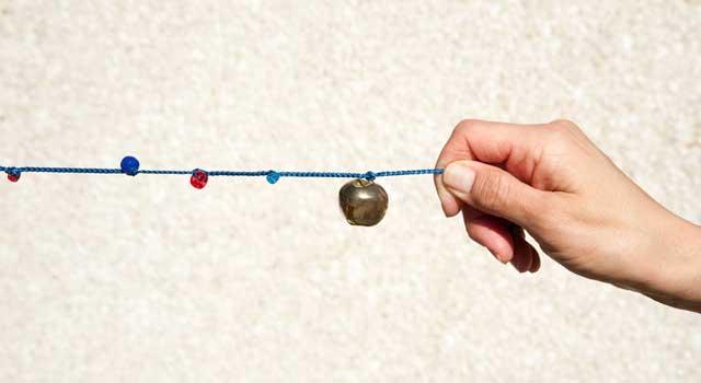 A hand is shown holding a blue string taut. A circular golden bell is at the right end of the string and four smaller beads are dispersed to the left of the bell.