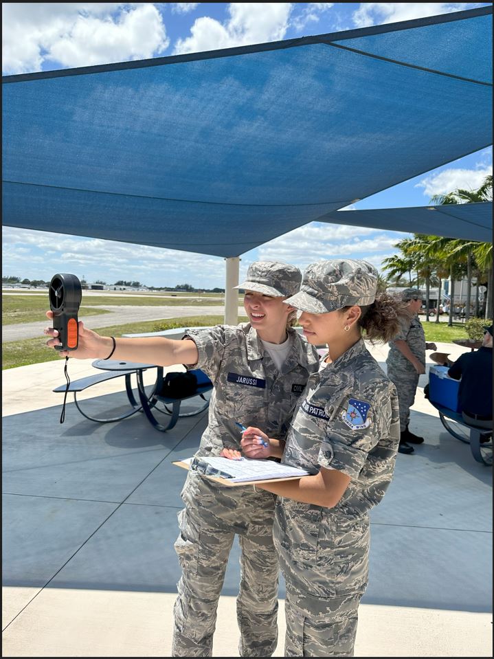 Two females cadets collecting wind measurements with clouds and palm trees in the background.