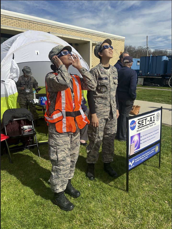 A female and male cadet stand in front of a tent staring at the sky while wearing solar viewing glasses.